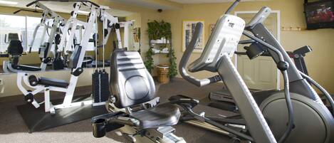 A gym with various exercise machines, a television on the wall, and a door in the background.