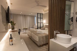 Una habitación de hotel moderna con una cama grande, una zona de estar, un baño con lavamanos espacioso y un televisor de pantalla plana montado en la pared.