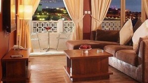 A modern living room with a wooden coffee table, a brown sofa, and a TV on the left. Large windows with curtains offer a view of the cityscape at night. - Spice Boutique Hotel (Salmiya)