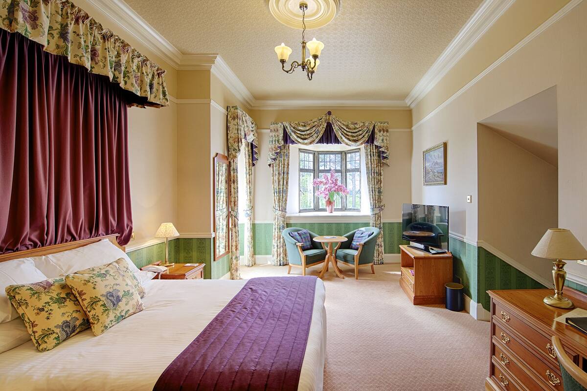 A well-appointed bedroom with a large bed, a desk, and a chair, featuring green and beige walls, floral curtains, and a chandelier.