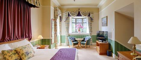 Un dormitorio elegante con una cama grande, un escritorio y una silla, paredes en tonos verde y beige, cortinas florales y una lámpara de araña.