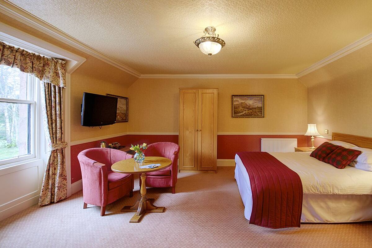 A hotel room with a bed, a bedside table with a lamp, a TV mounted on the wall, a wooden wardrobe, a small round table with a vase, and a window with curtains.