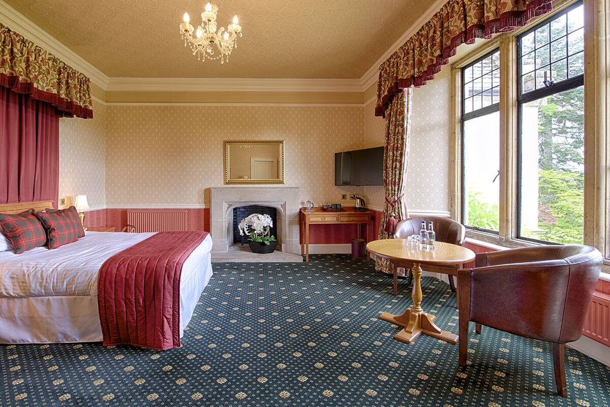 A hotel room with a large bed, a desk, a chair, a fireplace, and a large window.