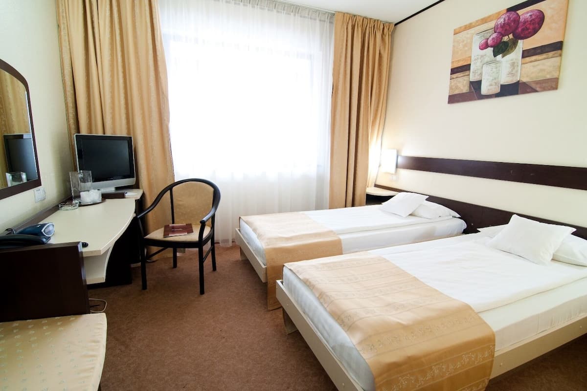 Ein Hotelzimmer mit zwei Einzelbetten, einem Schreibtisch mit Computer, einem Stuhl und einem Fenster mit Vorhängen.