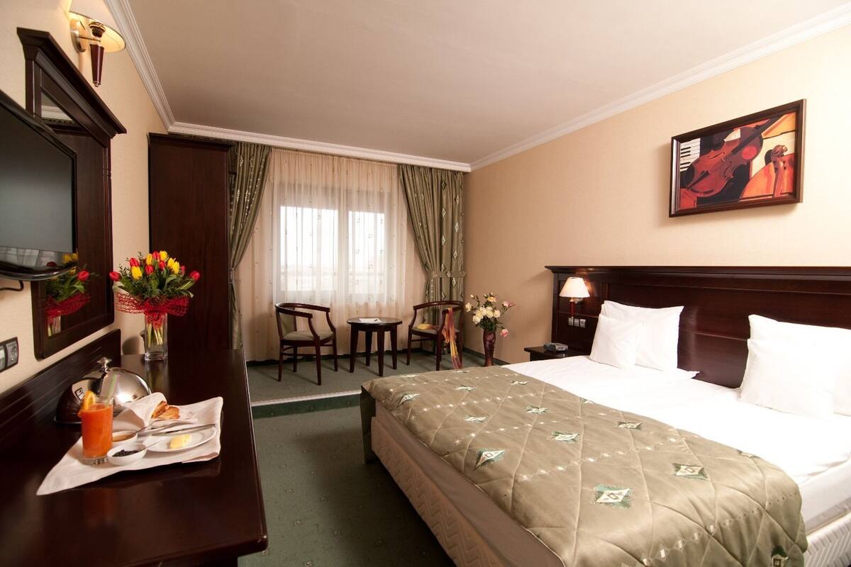 Ein Hotelzimmer mit einem Bett, einem Schreibtisch mit Stuhl, einem Fernseher, einem Bild an der Wand und einem Fenster mit Vorhängen.