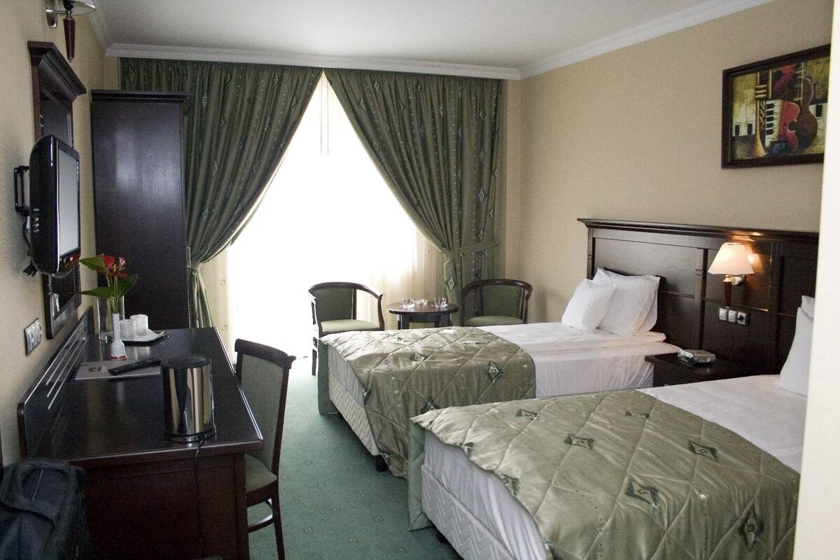 Ein Hotelzimmer mit zwei Betten, einem Schreibtisch mit Computer, einem Sessel, einem kleinen Tisch und einem Fenster mit Vorhängen.