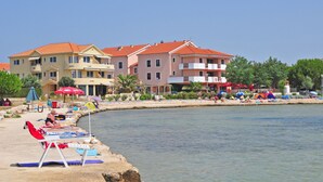 A waterfront hotel with multiple balconies, a beach area with sun loungers, and a clear view of the water.