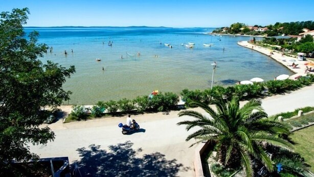 A coastal area with a clear view of the sea, a few boats, and a road with a motorcycle.