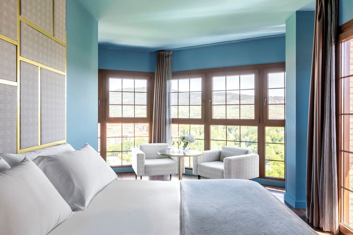 Habitación de hotel con una cama grande, dos sillones, una mesita con un florero y amplias ventanas con cortinas.