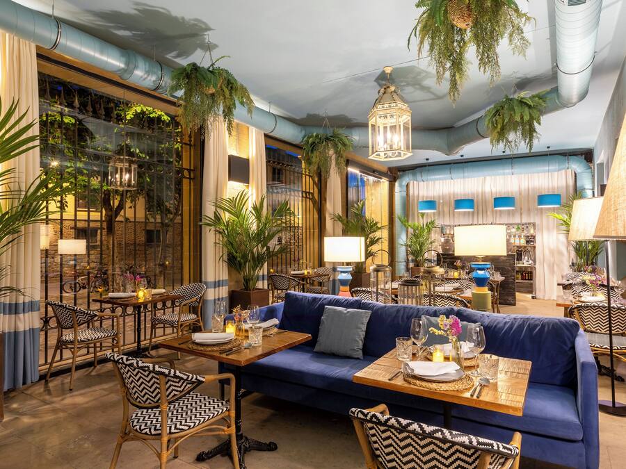 A modern interior with a blue sofa, patterned chairs, and a dining area with tables set for two.
