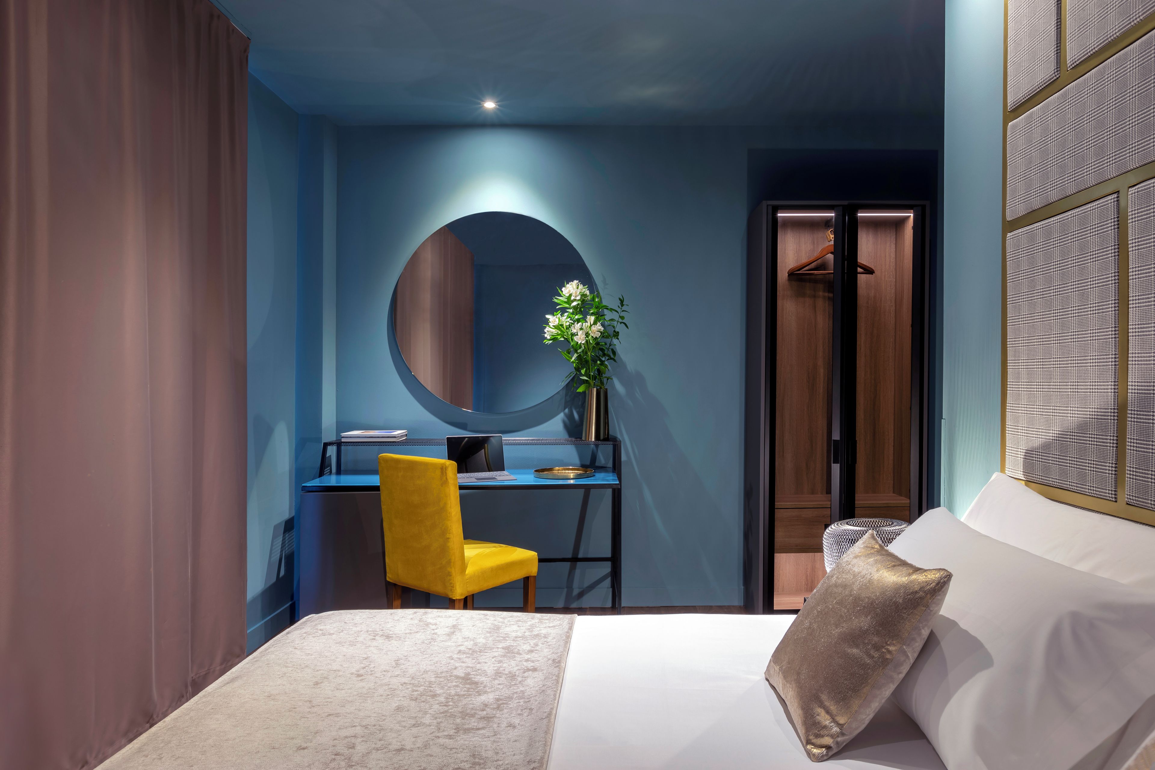 A modern bedroom with a bed, a desk, a chair, a round mirror, and a hanging plant.