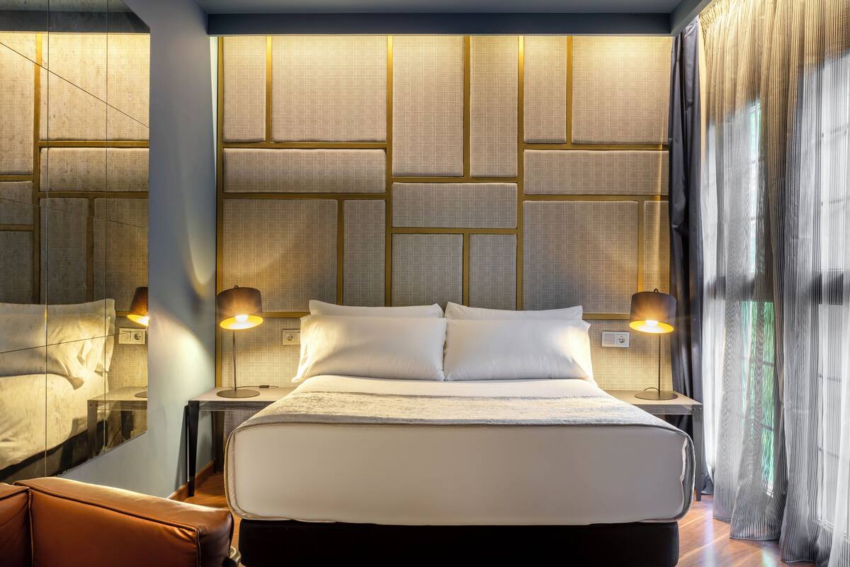 Una habitación de hotel moderna con una cama grande, dos lámparas de noche, un espejo y vistas al exterior a través de cortinas translúcidas.