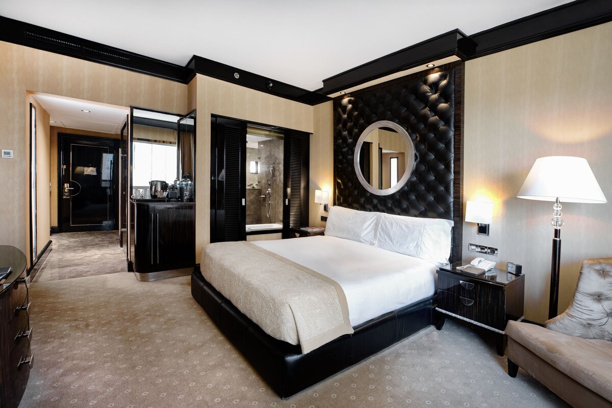 Habitación de hotel con una cama grande, una mesita de noche con lámpara, un espejo y vistas al baño.
