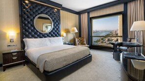 A hotel room with a large bed, a desk, a chair, a view of the city, and a large window.