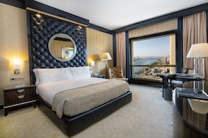 Habitación de hotel con una cama grande, un escritorio, una silla, vista a la ciudad y un ventanal amplio.