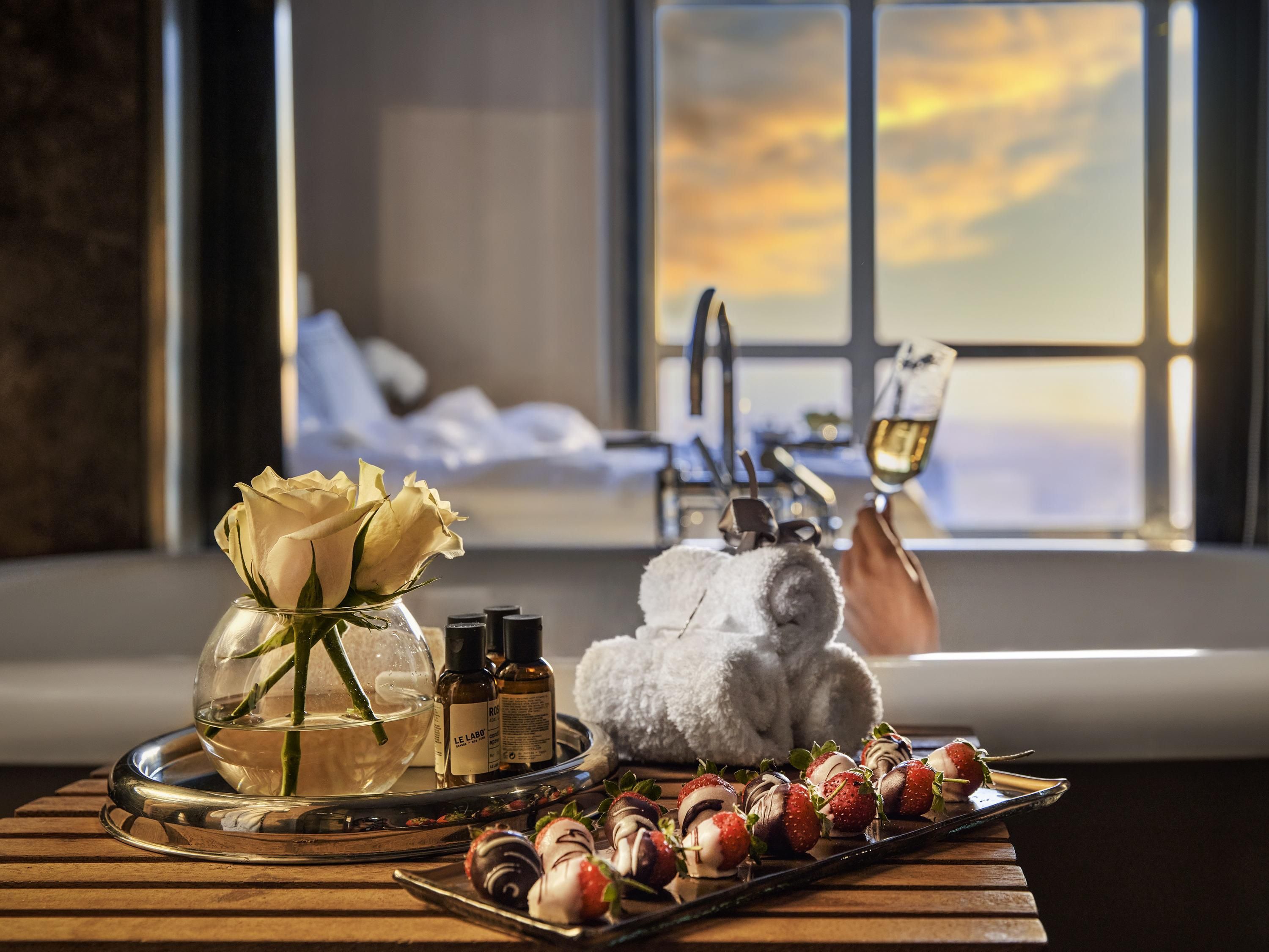 A room with a large window, a bed, a tray with strawberries, and a vase with a rose.