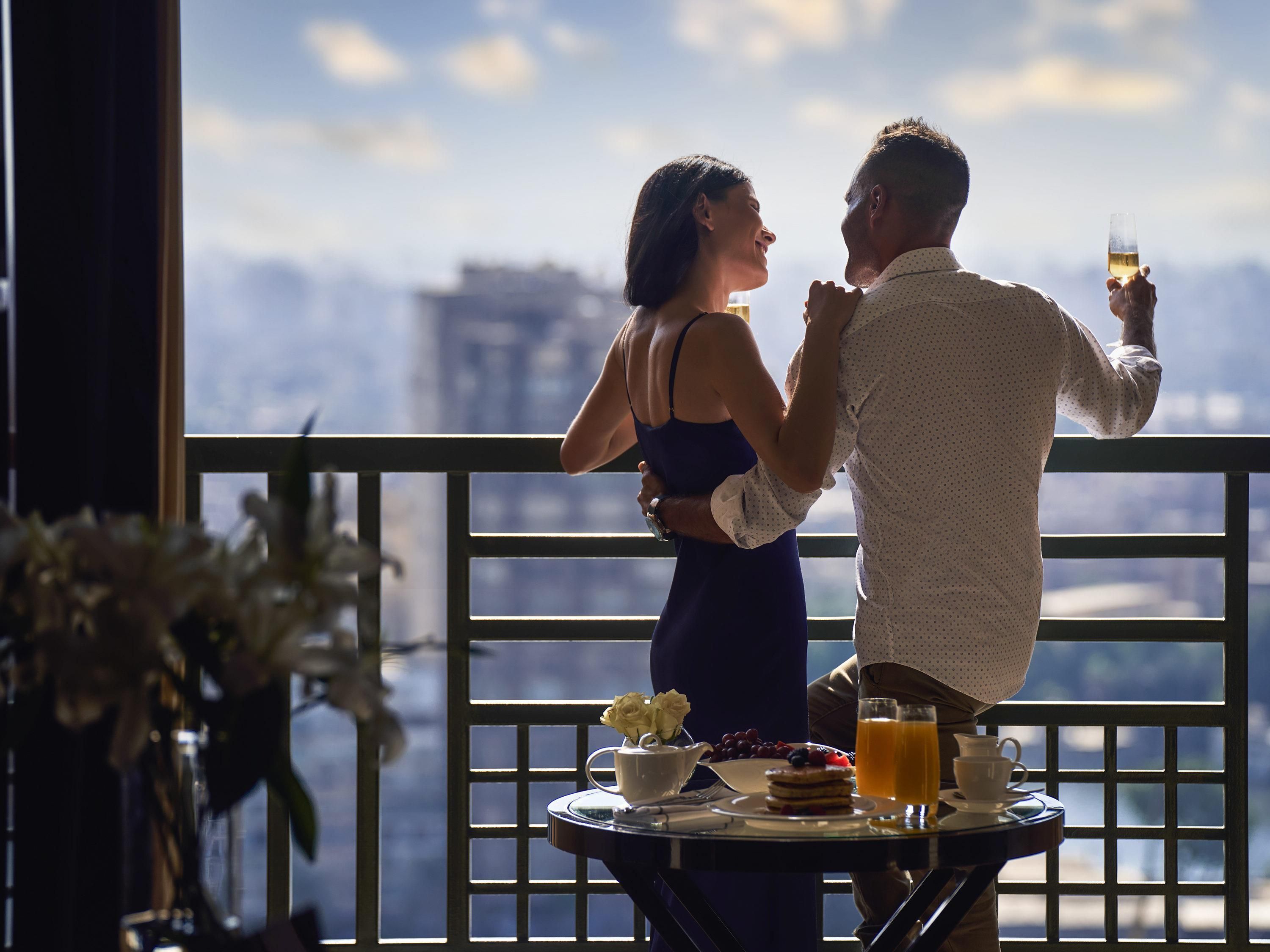 A couple dining at a table with a city view, a flower vase, and breakfast items.
