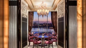 A dining area with a view of a cityscape at dusk, featuring a chandelier, ornate walls, and a patterned carpet. - Fairmont Nile City, Cairo (Cairo)