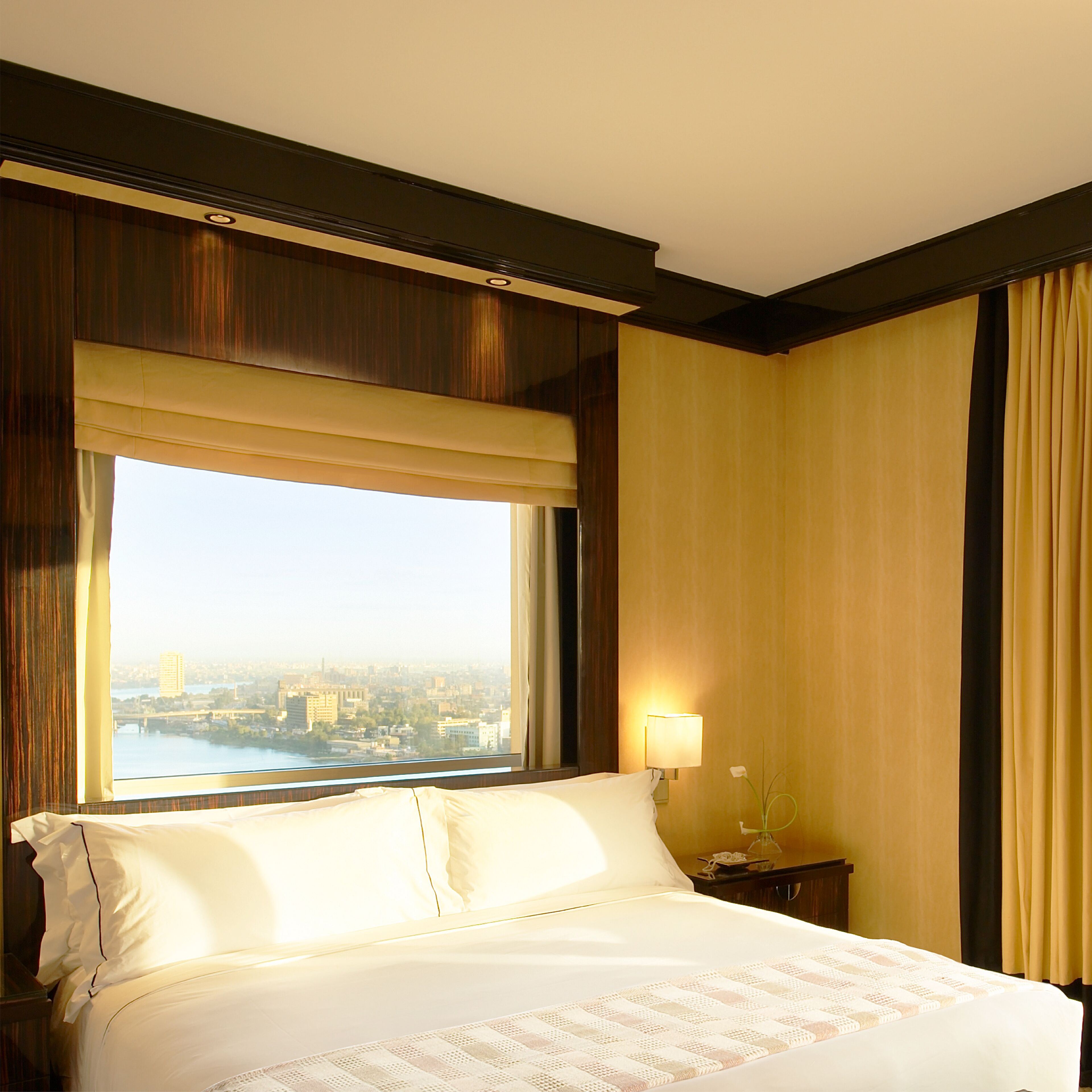 Une chambre d’hôtel avec un grand lit, une table de chevet avec une lampe et une vue sur le paysage urbain par une fenêtre avec des rideaux.