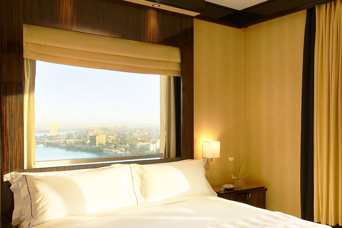 Habitación de hotel con una cama grande, una mesita de noche con lámpara y vistas a la ciudad a través de una ventana con cortinas.