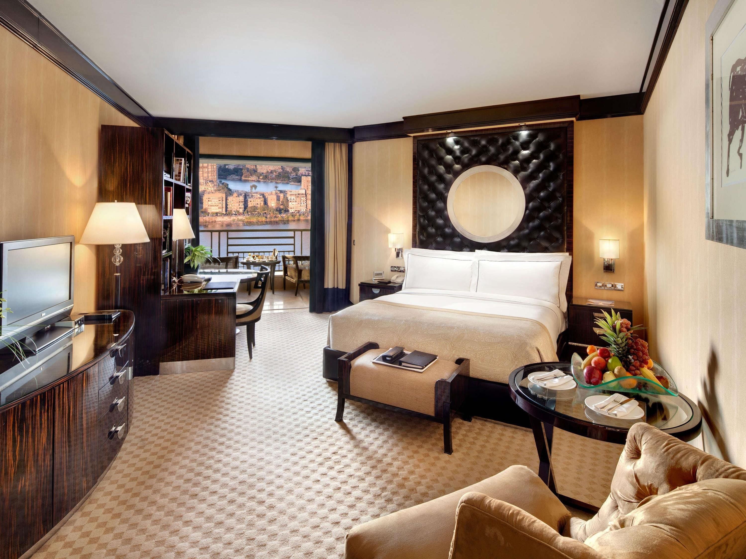 A hotel room with a bed, a desk, a TV, a dining area, and a view of a cityscape.