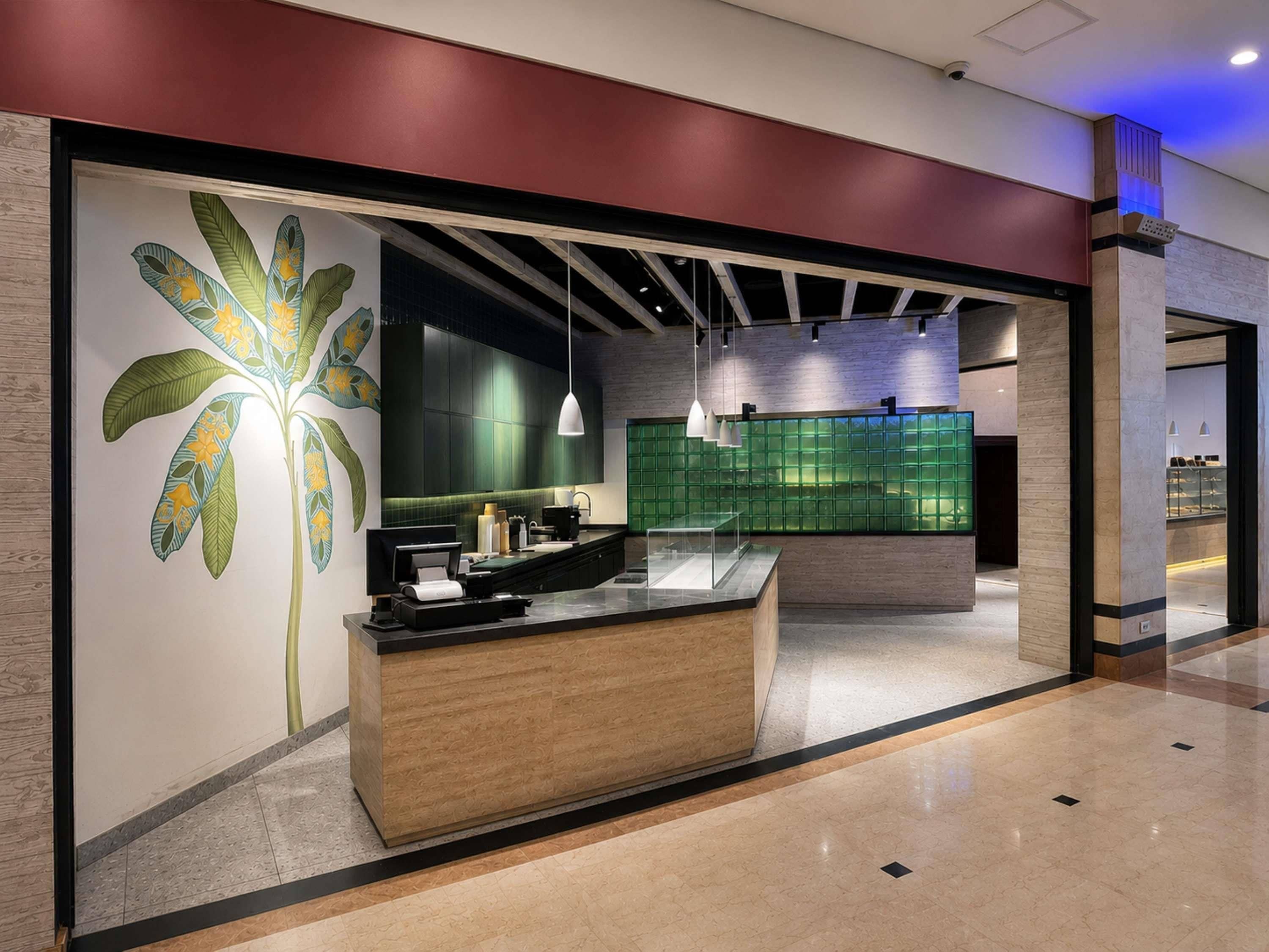 A modern reception desk with a wooden finish, a large decorative wall with a palm tree, and a green-tiled wall in the background.
