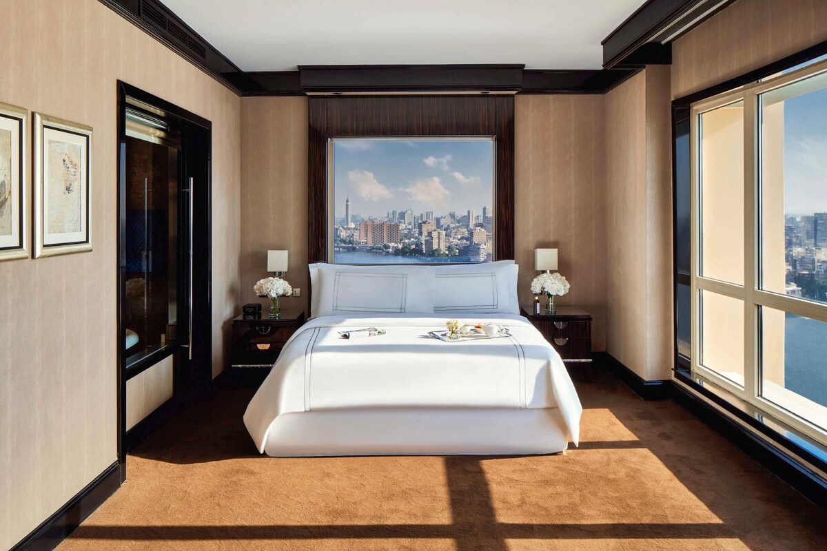 A hotel room with a large bed, two bedside tables with lamps, a large window with a city view, and framed artwork on the walls.