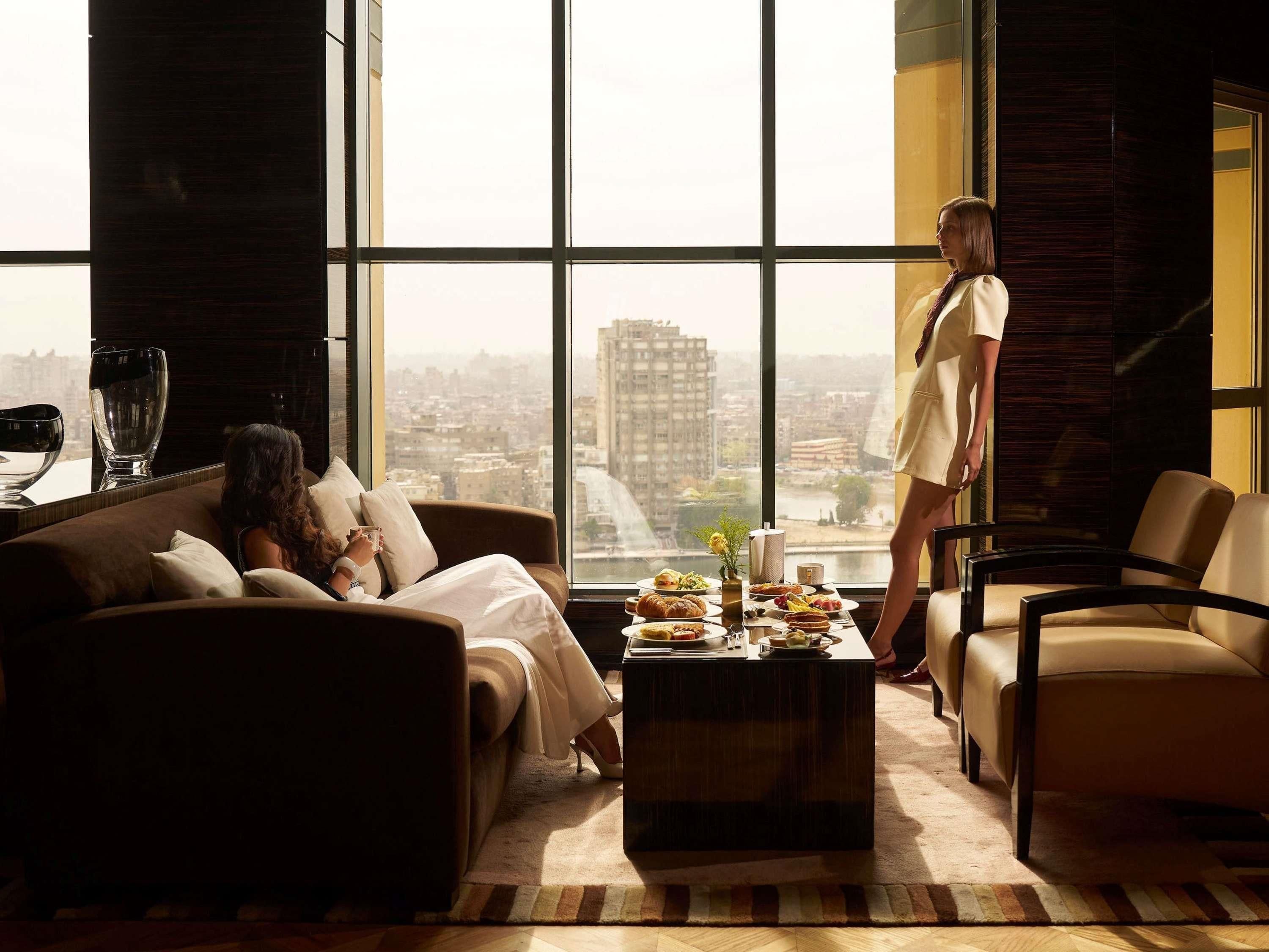 A modern living room with a large window, a sofa, a coffee table with food, and a city view.