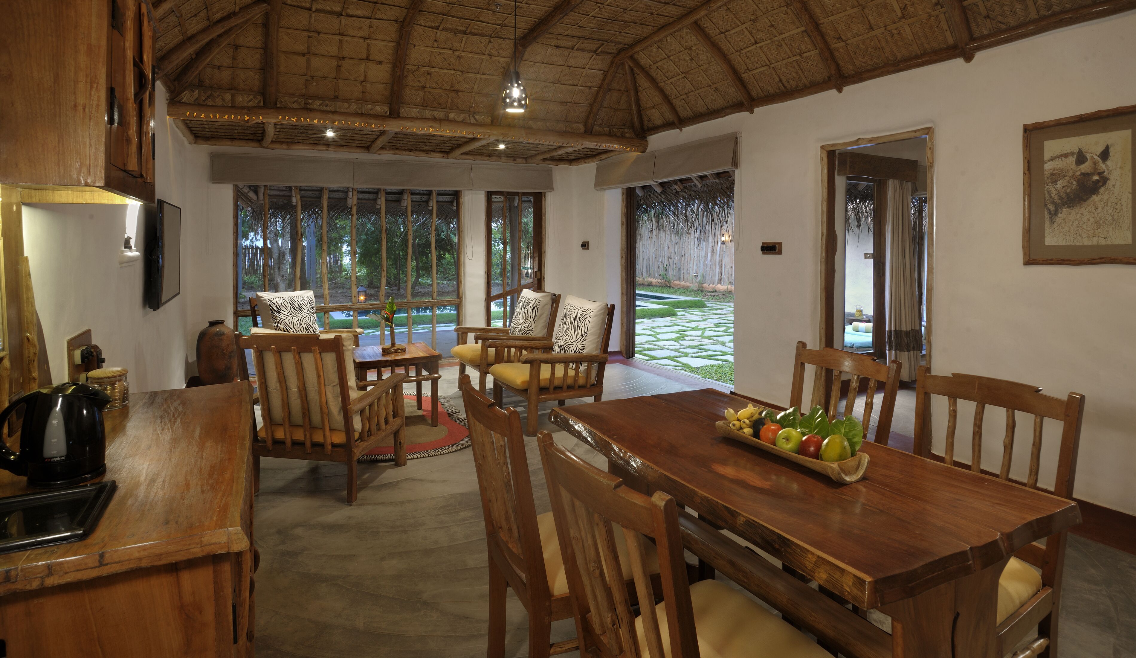 Un coin repas en bois, avec une table dressée pour quatre personnes, entourée de chaises. On peut voir un panier de fruits sur la table, et la pièce est dotée d’un plafond en chaume.