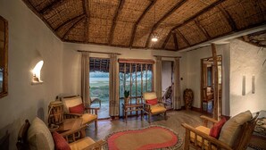 A spacious room with a high thatched ceiling, wooden furniture, and a view through a large window. - Evolve Back, Kabini (Heggadadevankote)