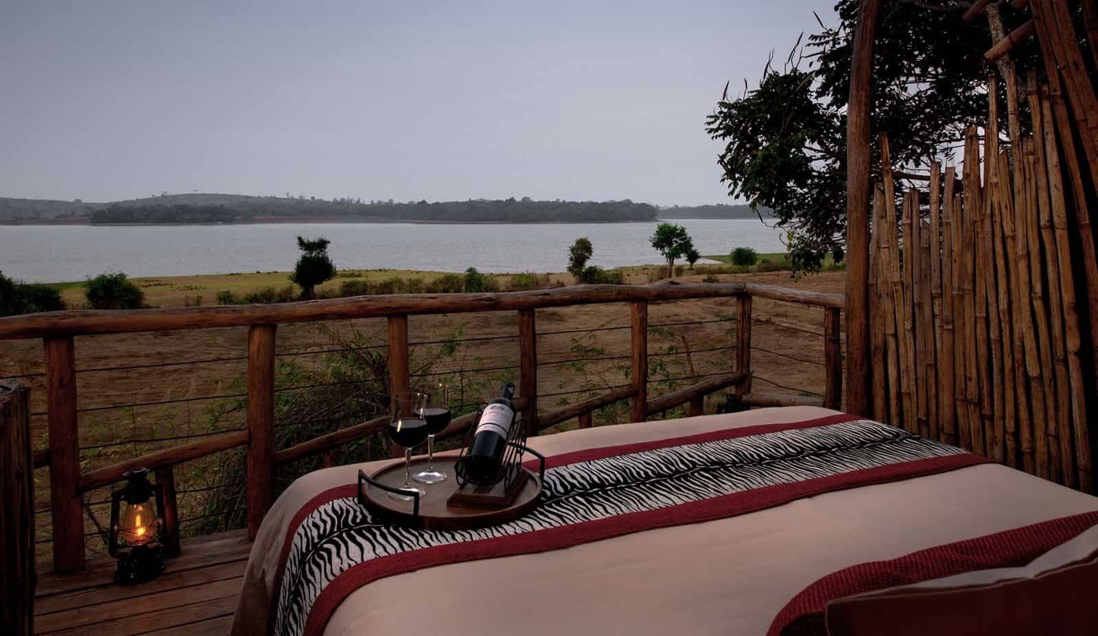 A bed with a zebra-patterned blanket, a bottle of wine, and a glass of red wine on a wooden deck overlooking a lake.
