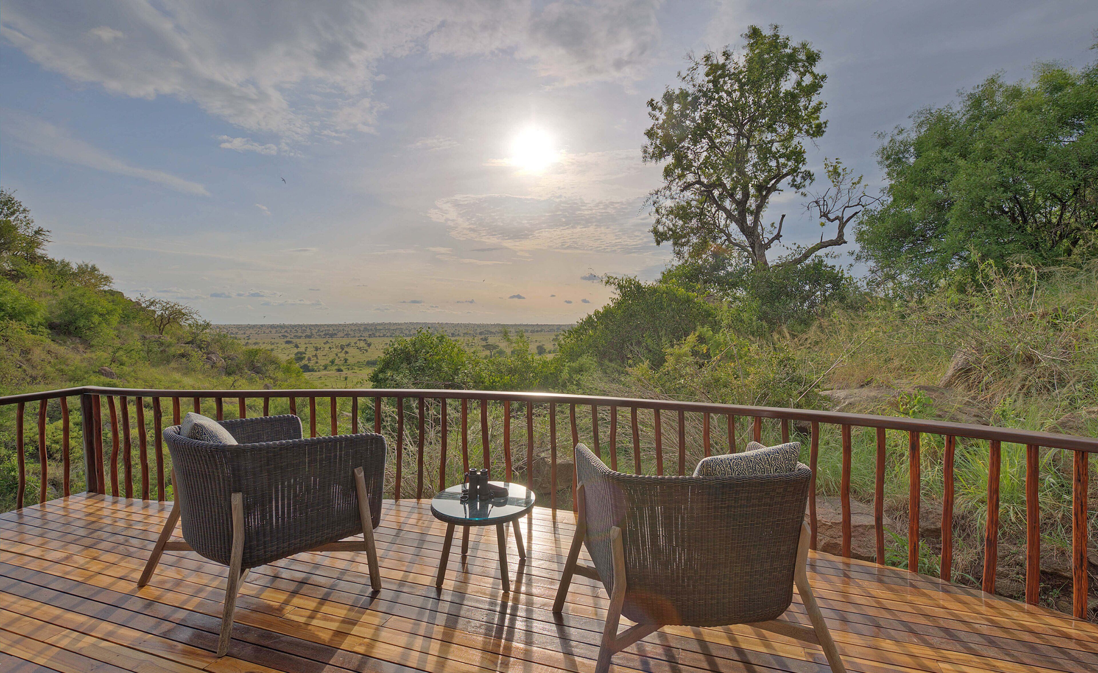 A wooden deck with wicker chairs and a glass table, overlooking a scenic landscape with trees and a distant horizon.