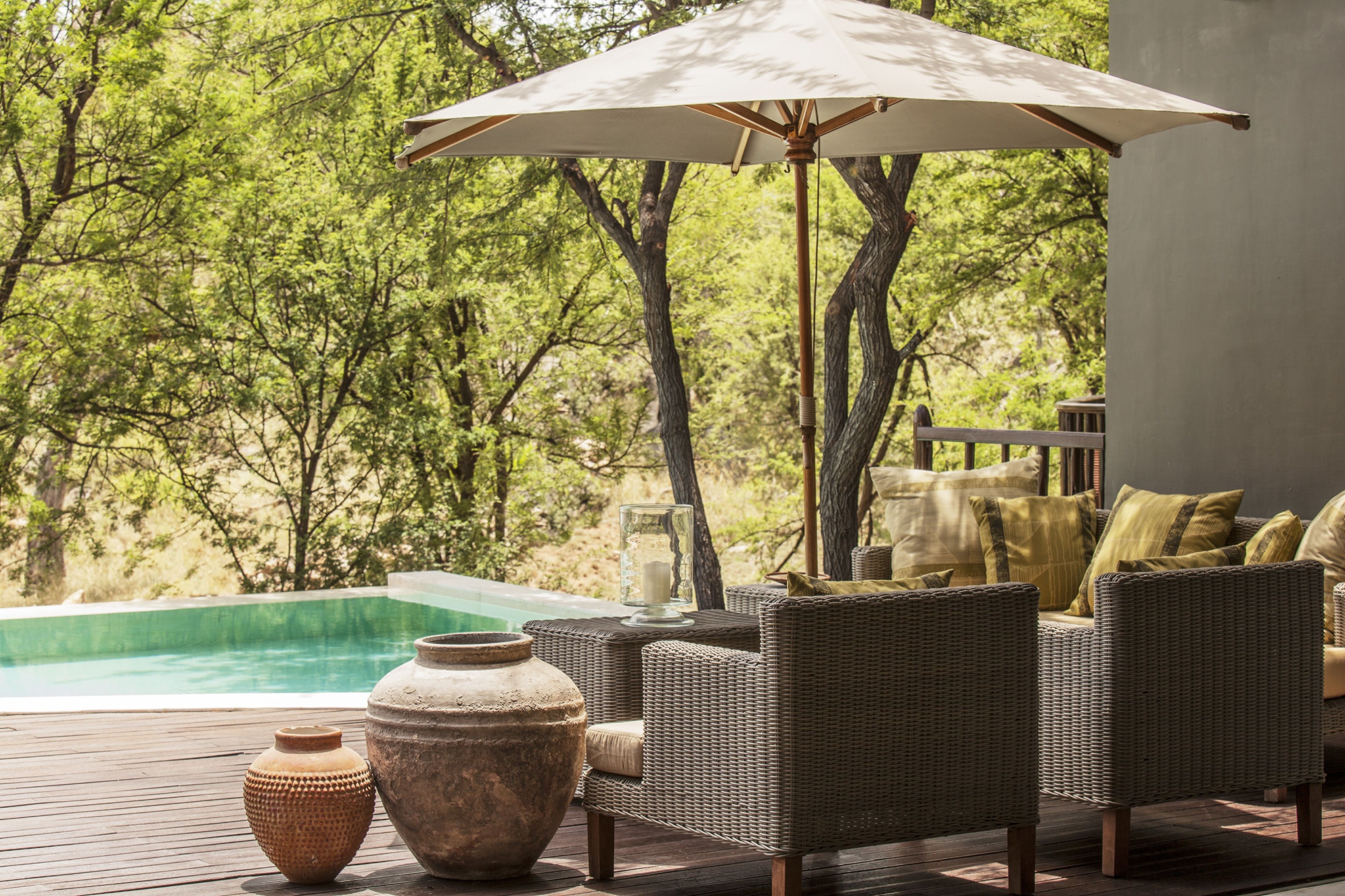 A deck with wicker furniture, a large umbrella, and a view of a swimming pool surrounded by trees.