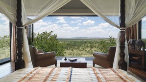 A room with a large window, two leather chairs, a wooden coffee table, and a view of a landscape with trees and mountains.