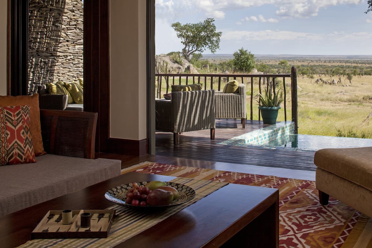 Ein Wohnzimmer mit einer Couch, einem Couchtisch mit Früchten und einem Brettspiel, ein Balkon mit Korbmöbeln und Blick auf die Savanne.