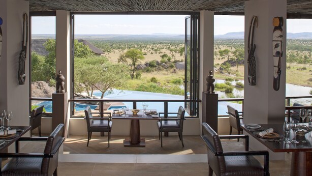 A dining area with tables and chairs, large windows offering a view of a natural landscape.
