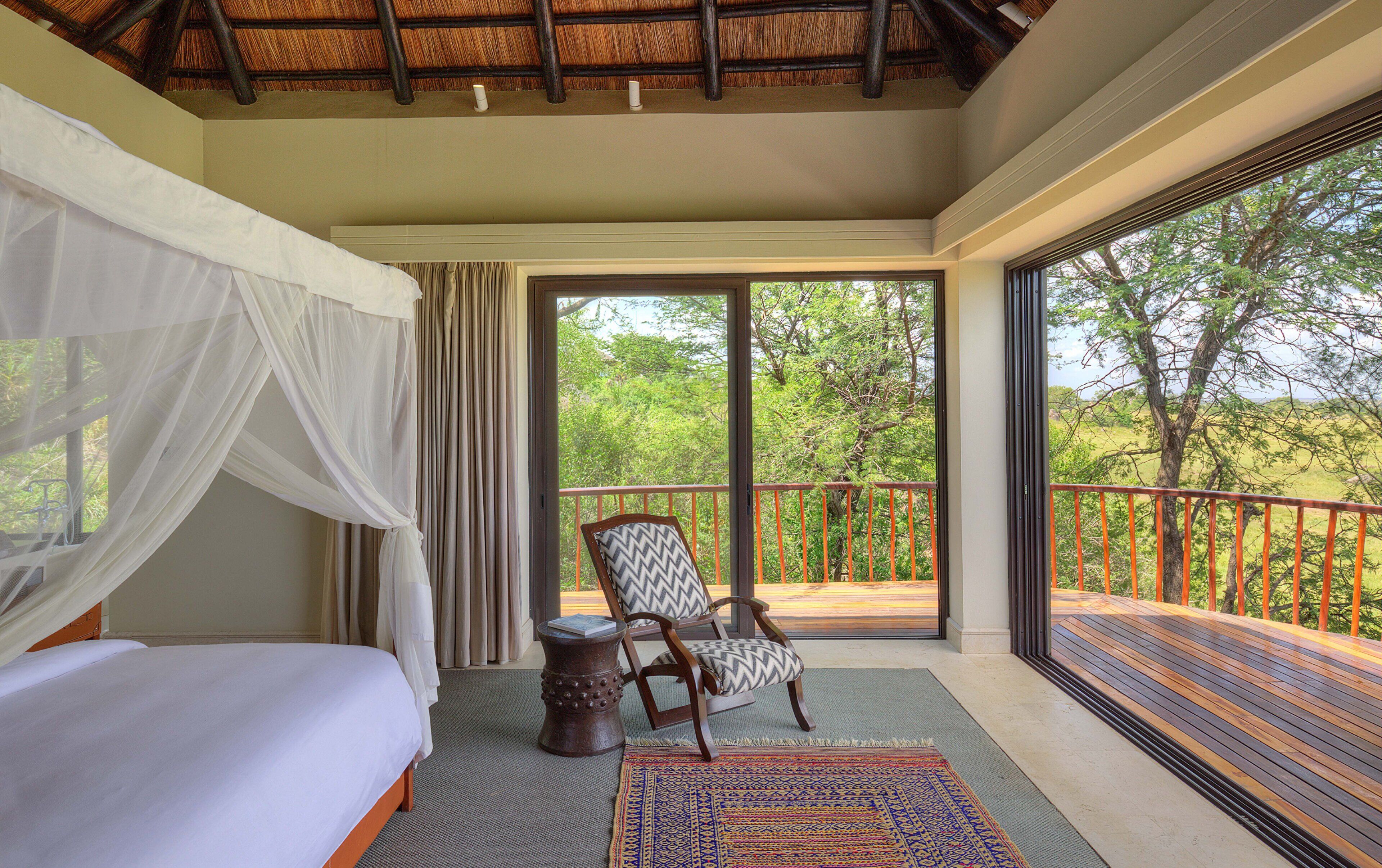 A bedroom with a bed, a chair, a balcony with a railing, and a view of trees.
