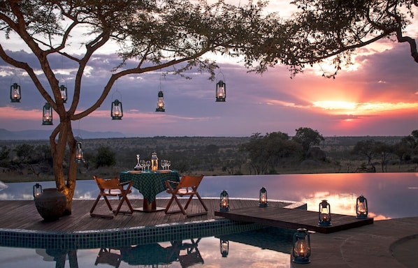 A serene outdoor dining area with a table set for two, overlooking a calm body of water and a sunset.