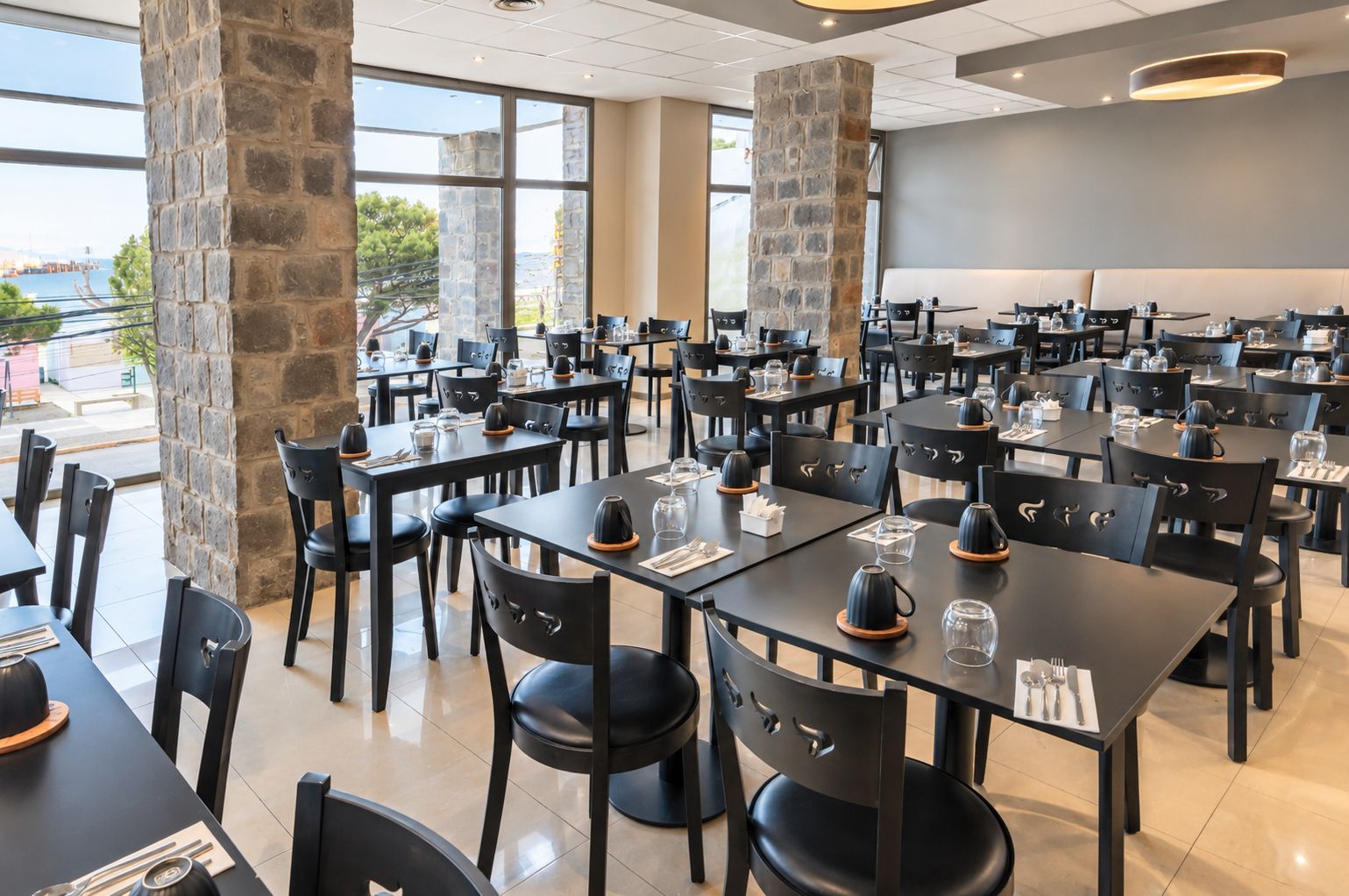 Un restaurante espacioso con varias mesas y sillas, pilares de piedra y amplias ventanas con vistas al exterior.
