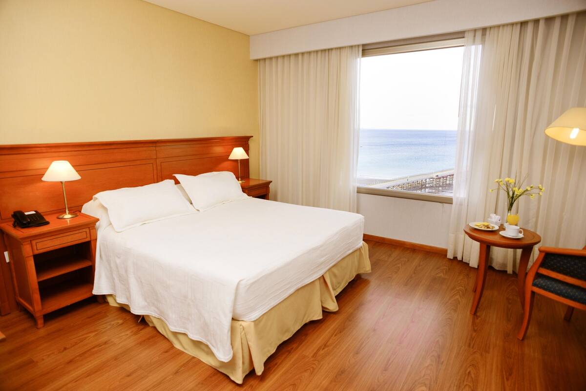 Une chambre d’hôtel avec un grand lit, une tête de lit en bois, des lampes de chevet, une petite table avec un vase et une vue sur la mer.