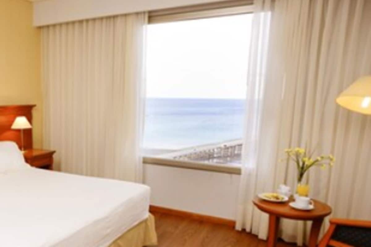 A hotel room with a bed, a nightstand, a lamp, a window with a view of the sea, and a small table with a vase of flowers.
