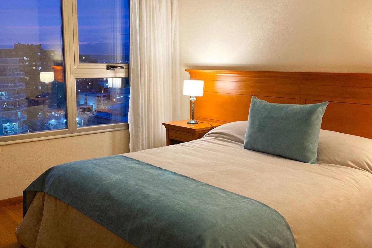 Une chambre d’hôtel avec un grand lit, une table de chevet avec une lampe, une vue sur la ville par la fenêtre et une tête de lit en bois.