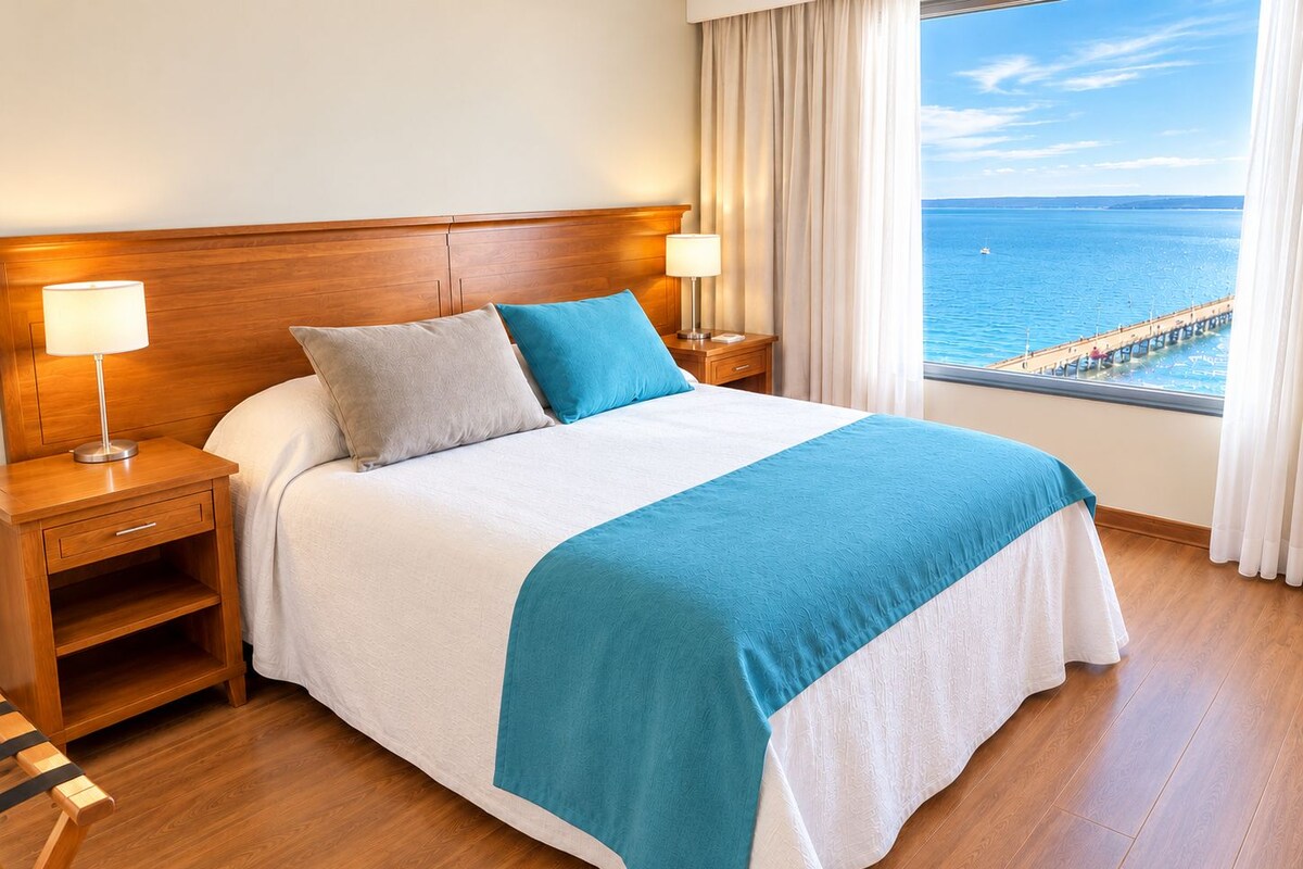 A hotel room with a large bed, wooden headboard, two bedside tables with lamps, a view of the sea through a window, and a wooden floor.