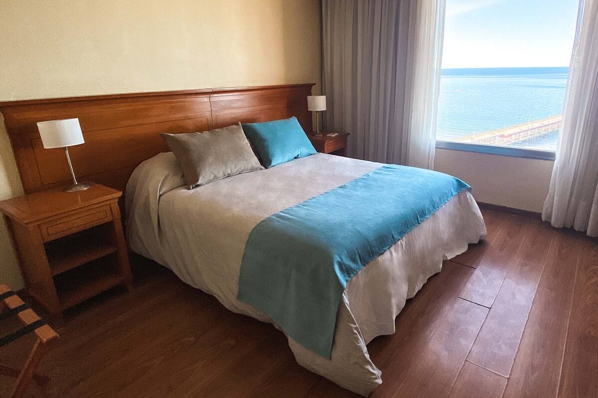 Une chambre d’hôtel avec une tête de lit en bois, un lit recouvert d’une courtepointe bleue et blanche, deux tables de chevet avec des lampes, et une vue sur la mer par la fenêtre.