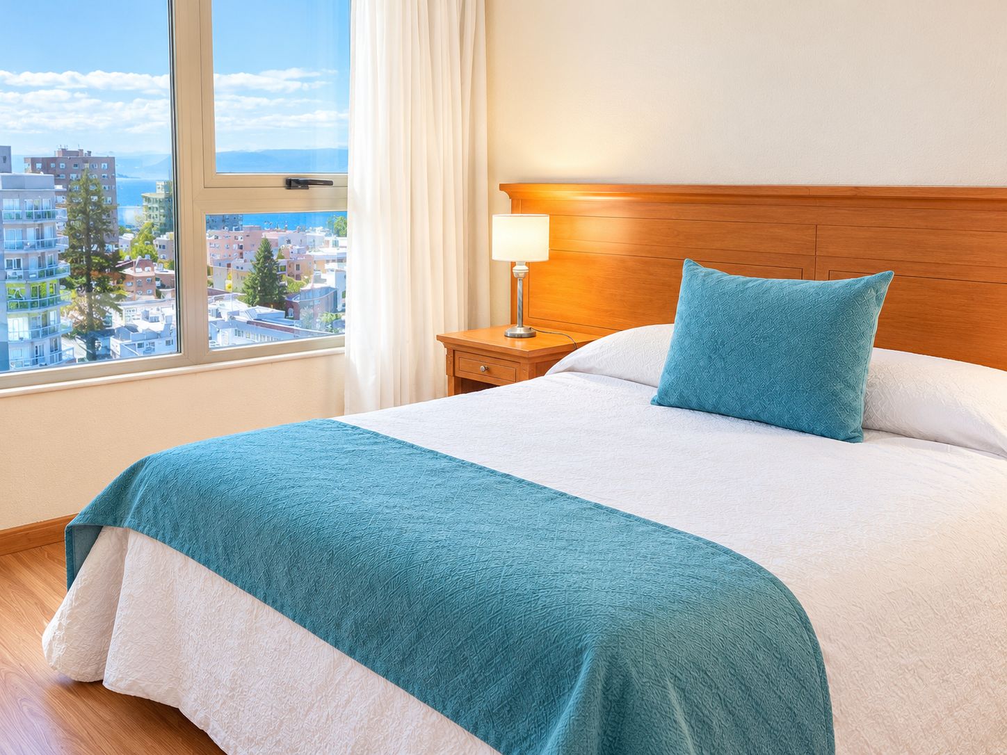 A hotel room with a large bed, a nightstand with a lamp, a view of the city through the window, and a wooden headboard.