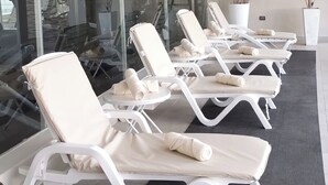 Una hilera de sillas de descanso blancas con toallas, sobre un piso de baldosas con un tapete negro, en un ambiente interior moderno con paredes de vidrio y plantas en macetas.