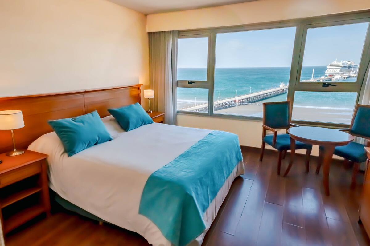 Une chambre d’hôtel avec un grand lit, un bureau avec une chaise et une vue sur l’océan et la jetée.