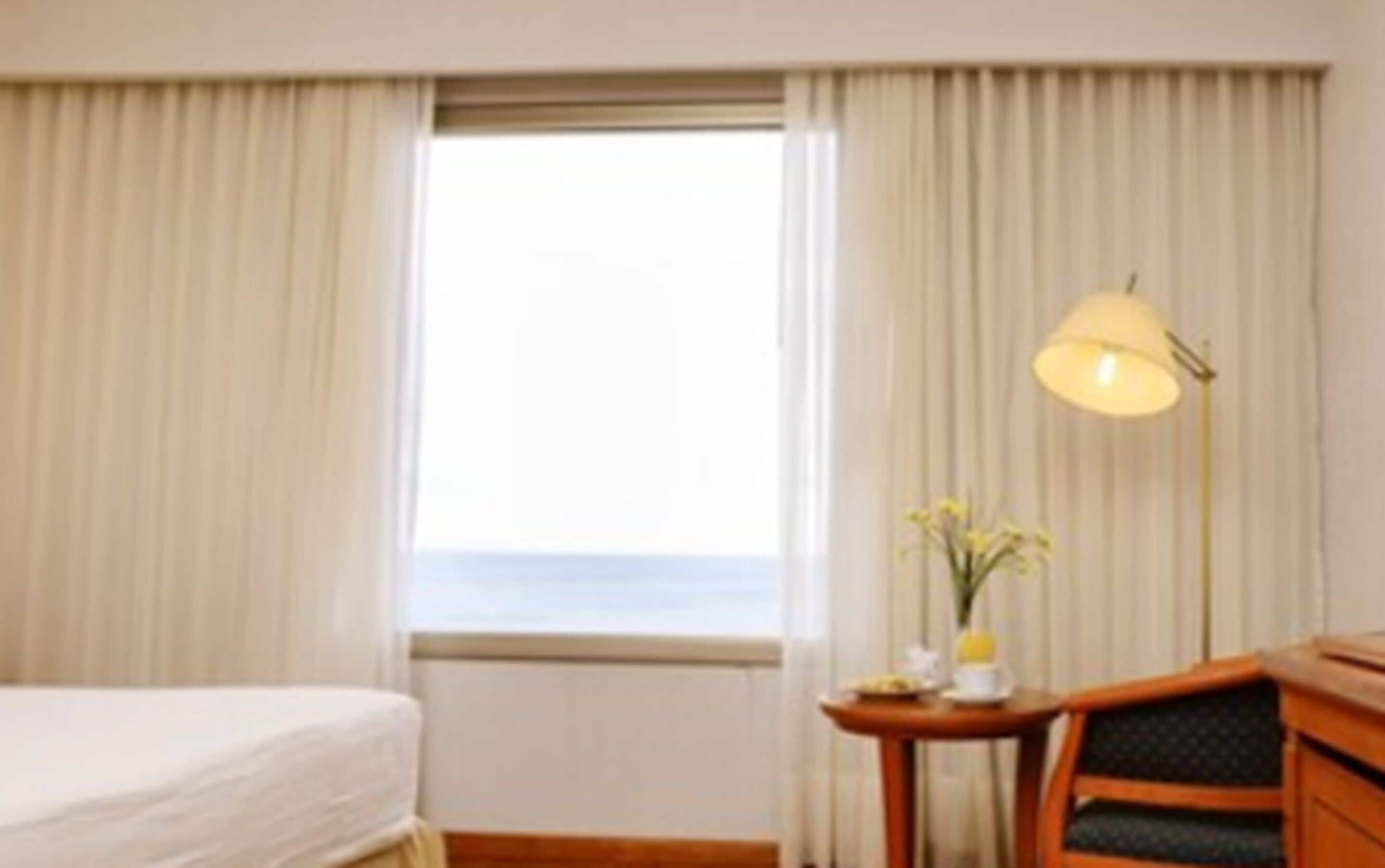 A hotel room with a bed, a small table with a lamp, and a view of the sea through the window.