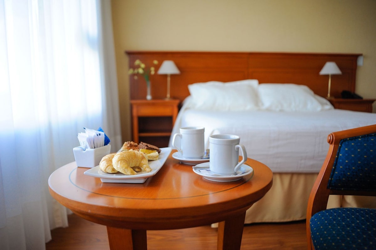 A hotel room with a bed, a bedside table with a lamp, a chair, and a breakfast table with croissants and coffee cups.