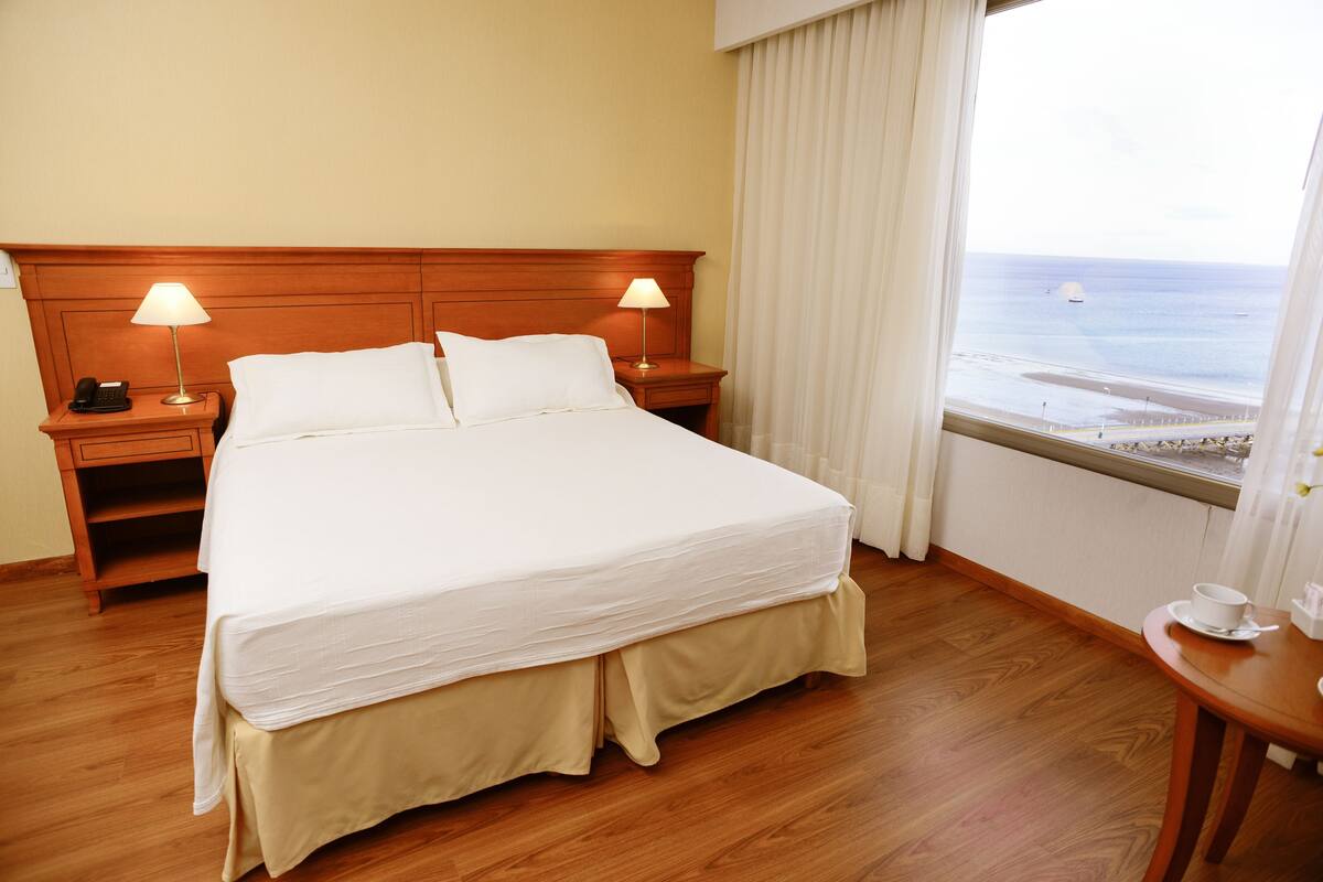 Une chambre d’hôtel avec un grand lit, deux tables de chevet avec des lampes, une vue sur la plage et une fenêtre avec des rideaux.